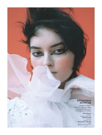 Close-up portrait of a woman with dark hair and dramatic eyeliner, wearing a white ruffled top against an orange backdrop.