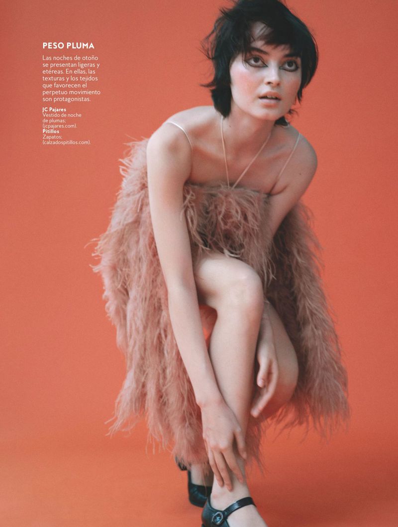 Model Greta Hajwos crouches in a blush feather dress and black shoes against a vibrant coral orange seamless studio background.