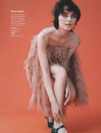 Model Greta Hajwos crouches in a blush feather dress and black shoes against a vibrant coral orange seamless studio background.