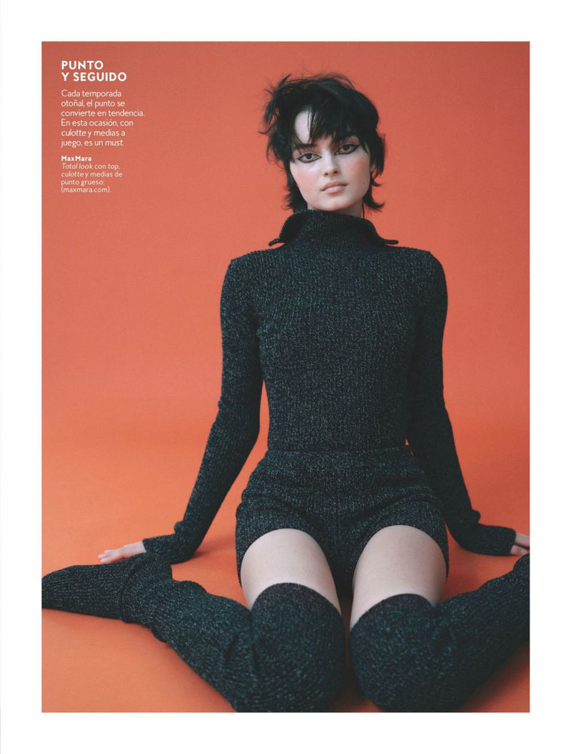 A young woman with dark hair poses in a dark, long-sleeved top and shorts, with matching thigh-high leg warmers, against an orange background.