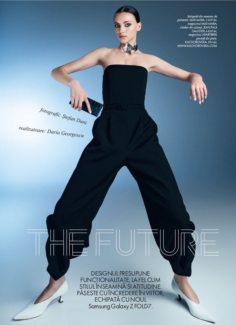 Fashion editorial shot of a model in a black strapless jumpsuit and white heels, holding a phone, against a gradient blue background.
