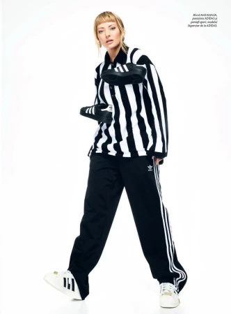 Full shot of a woman in a striped shirt and Adidas track pants posing with black sneakers against a white backdrop in a studio setting.