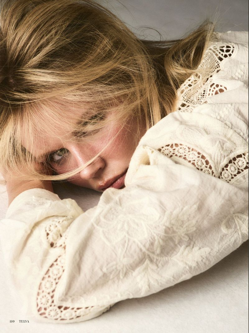 Close-up portrait of a young blonde adult woman in a white embroidered blouse, resting her head on her arms, with a neutral expression.