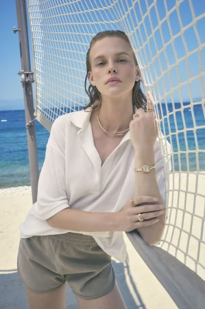A woman in casual clothing poses on a sandy beach with blue sky, ocean, and volleyball net in the background; wearing jewelry.