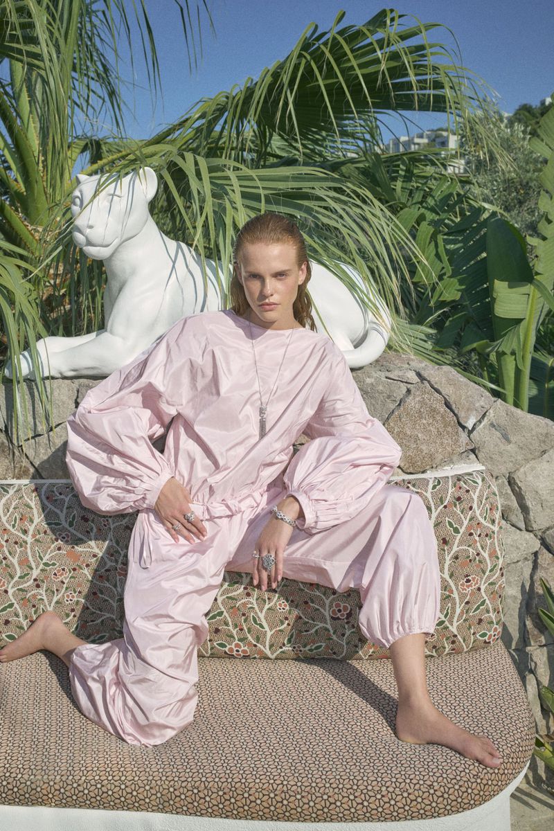 A fashion model sits on an outdoor couch in an oversized pink jumpsuit, accessorized with silver jewelry, with tropical foliage behind her.