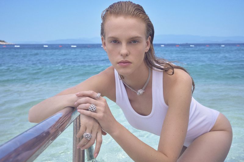 Attractive woman in a white swimsuit and jewelry leans on a metal bar in shallow, turquoise ocean water with a coast in the distance.