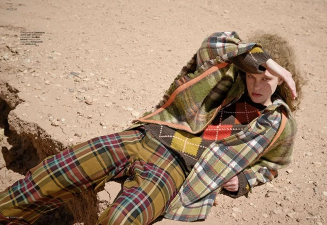 Fashion editorial shot of a model lying on arid ground wearing a plaid jacket and pants with an argyle sweater and curly hair.