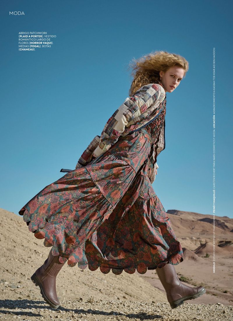 A young woman walks across a desert landscape wearing a long, floral dress, a patchwork jacket, and brown boots against a clear blue sky.