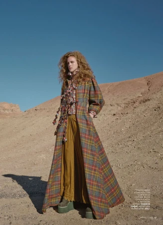 A young woman with red curly hair stands in a desert landscape wearing a long plaid coat, ruffled blouse, and mustard-colored pants.