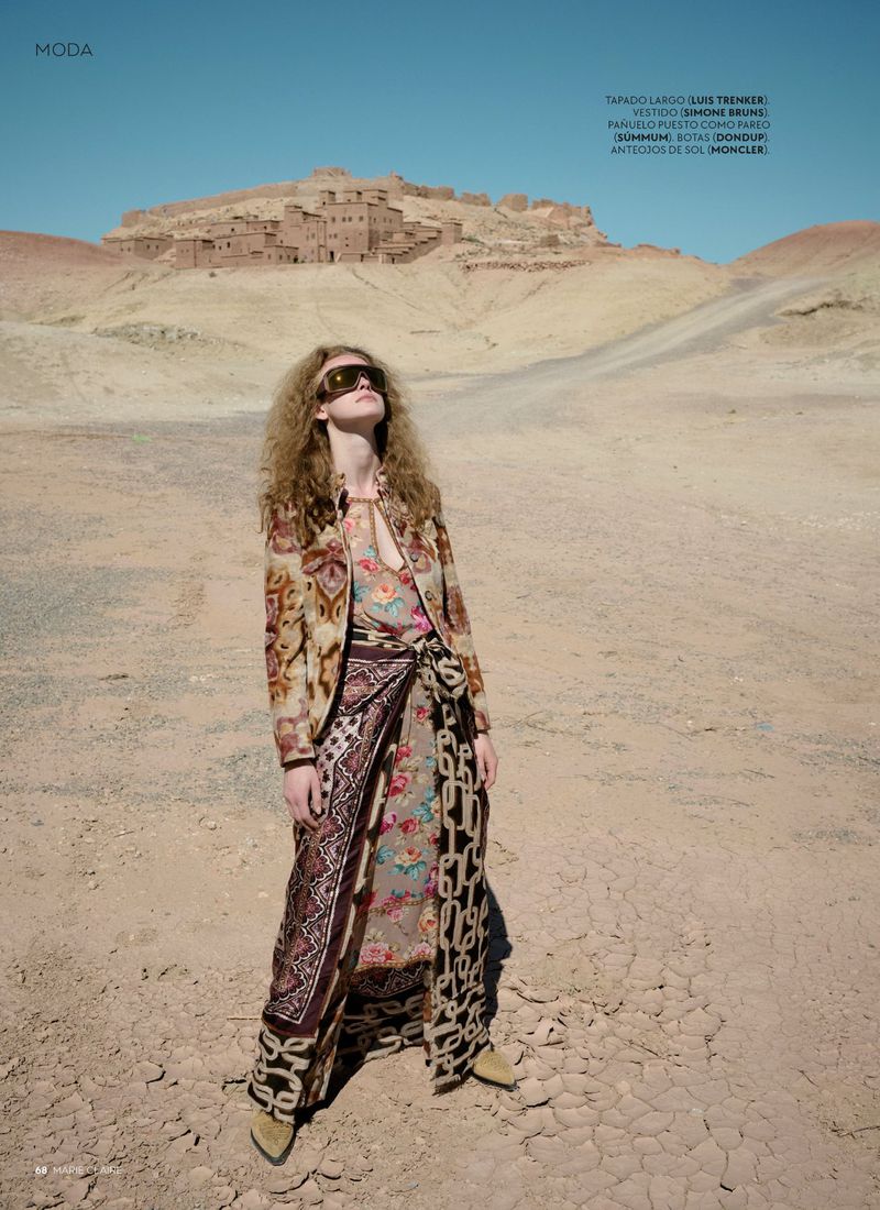 Fashion shot of a woman in a patterned jacket and skirt, standing in a desert landscape with a village in the background under a blue sky.