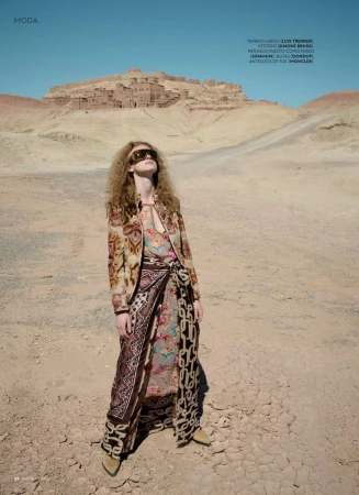Fashion shot of a woman in a patterned jacket and skirt, standing in a desert landscape with a village in the background under a blue sky.