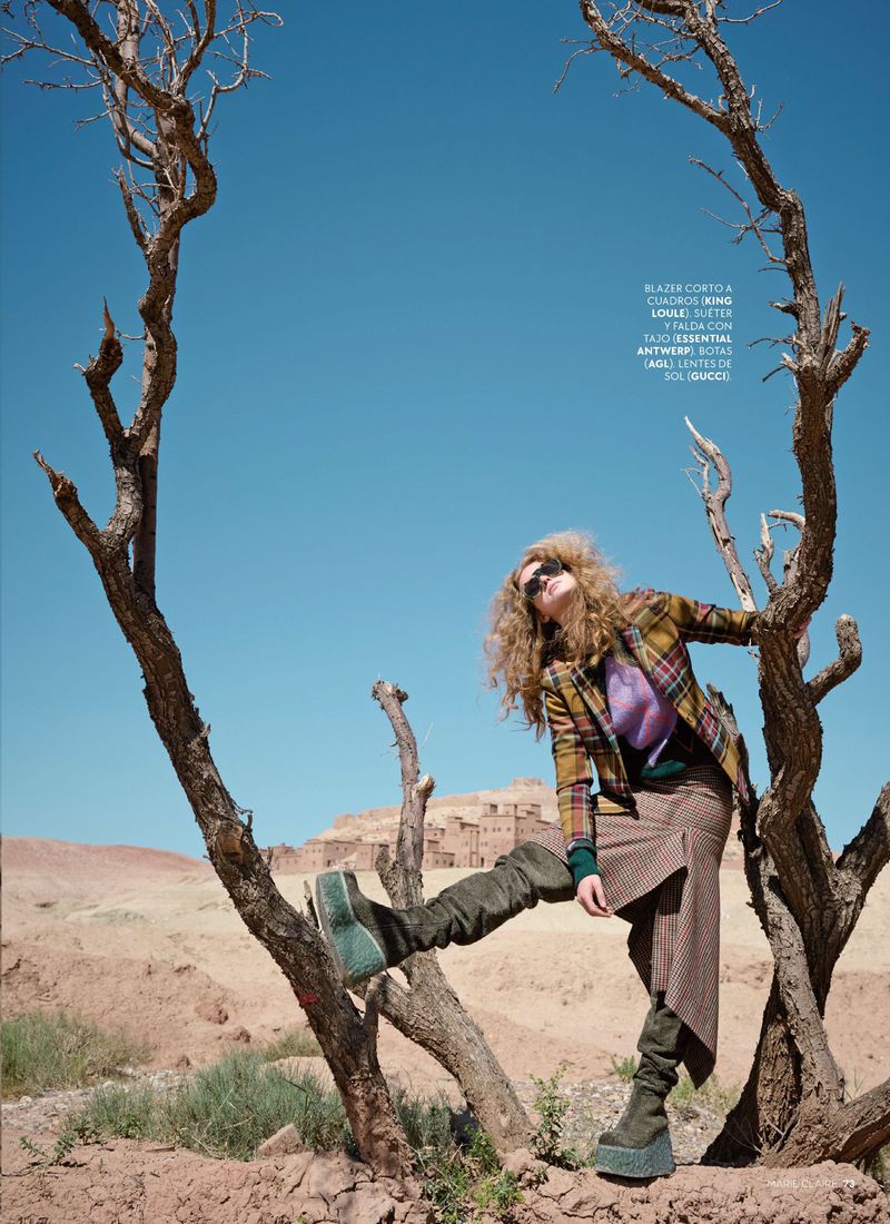 Fashion editorial shot of a curly-haired woman in a plaid jacket, skirt, and platform boots posing near a dead tree against a desert backdrop.