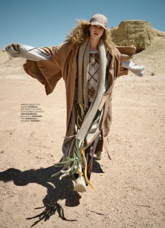 Fashion editorial featuring a fair-skinned adult woman in a tan coat, argyle sweater, and cap walking in an arid desert landscape.