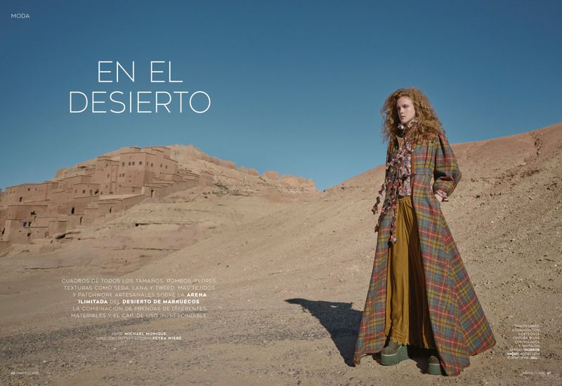Fashion editorial shot of a woman wearing a plaid coat and yellow pants in a desert landscape with a historic building backdrop.