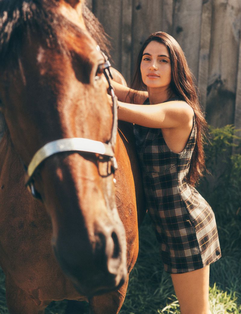 Woman in plaid mini dress poses with a brown horse in an outdoor, sunny, rural environment against a wood fence background.