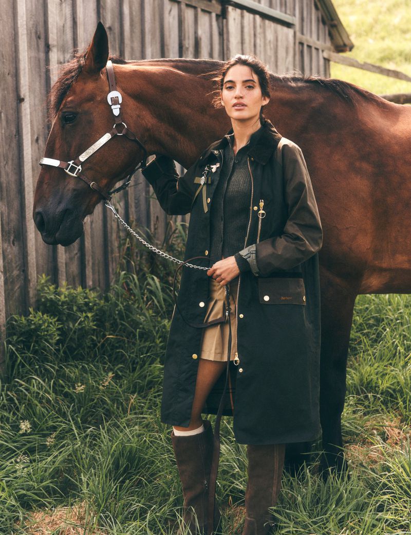 A chestnut-colored horse stands next to a woman outdoors in a long, dark coat, skirt, and boots, near a weathered wood barn.