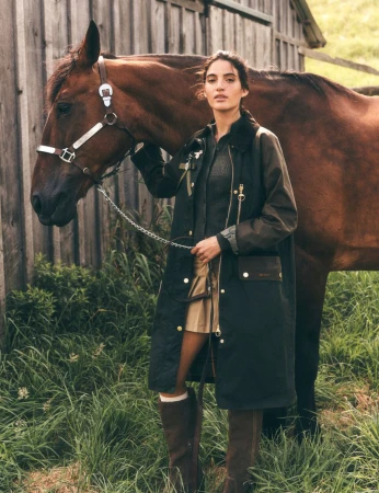 A chestnut-colored horse stands next to a woman outdoors in a long, dark coat, skirt, and boots, near a weathered wood barn.