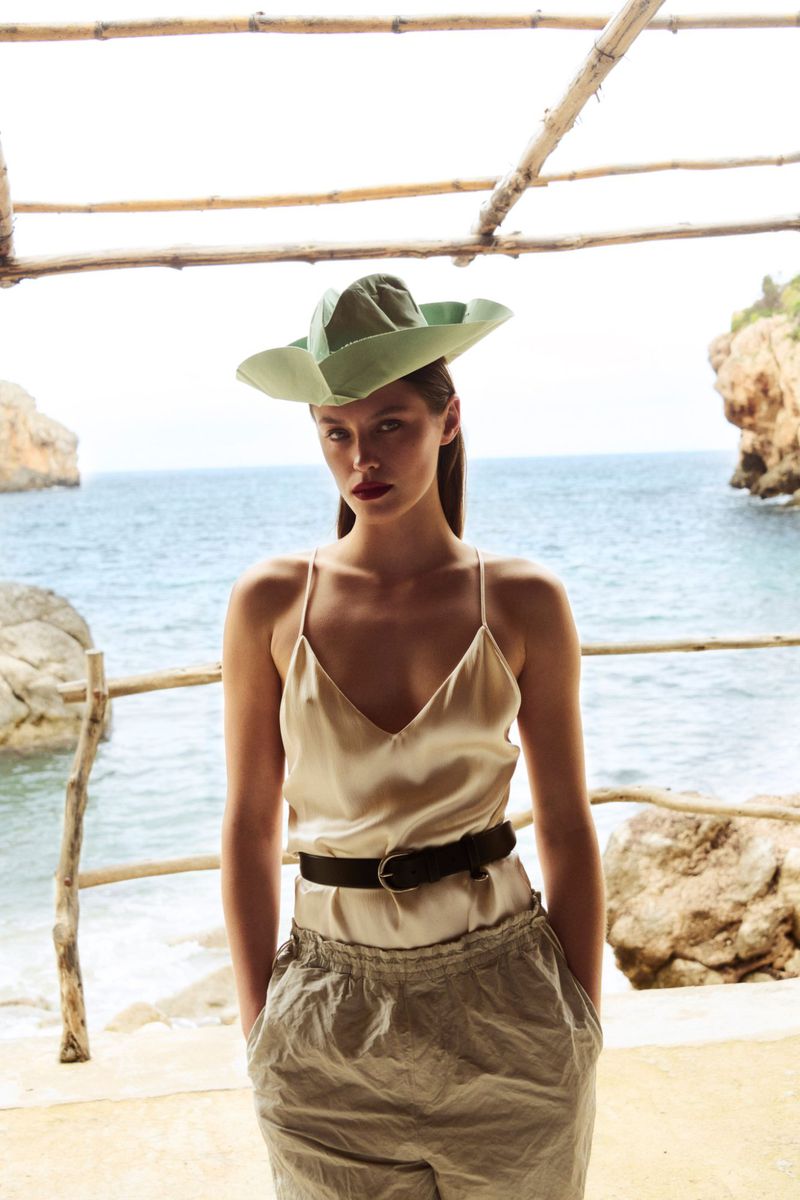 A woman in a green hat and beige outfit stands on a beach deck overlooking the ocean, with rocks and cliffs in the background.