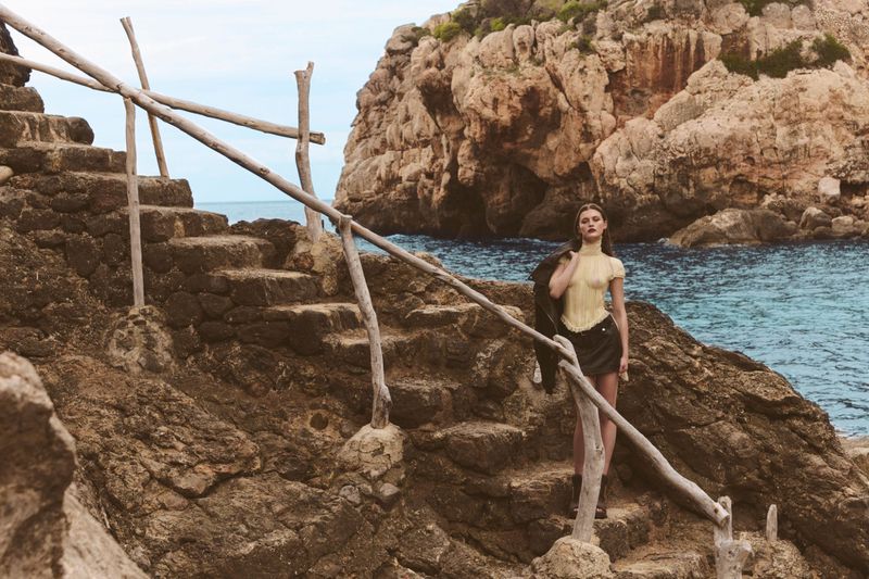Fashionable woman in yellow top and black skirt stands on stone steps with wood railing near the sea, wearing jacket over her shoulder.