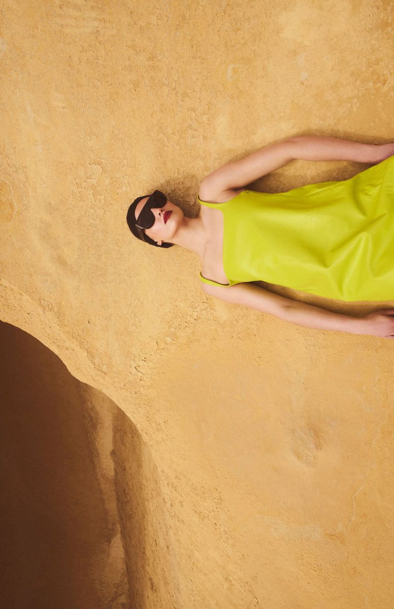 Overhead shot of a stylish woman in a neon dress and sunglasses lying on a textured tan surface, fashion editorial, studio lighting.