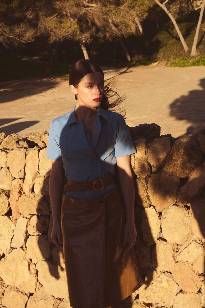 A woman poses outdoors in front of a stone wall, wearing a blue top and brown wrap skirt, with trees and light in the background.