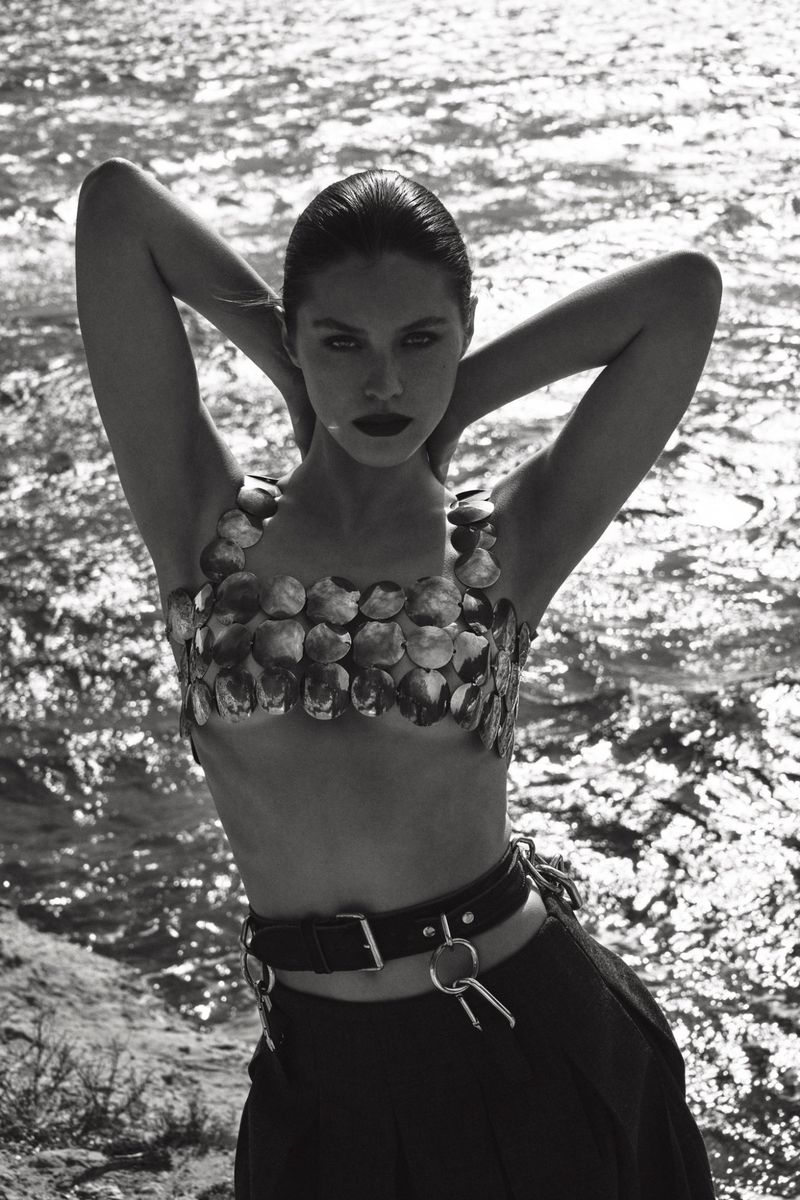 Monochrome portrait of a woman in a shell top and wide-leg pants posing by a body of water with her hands behind her head.
