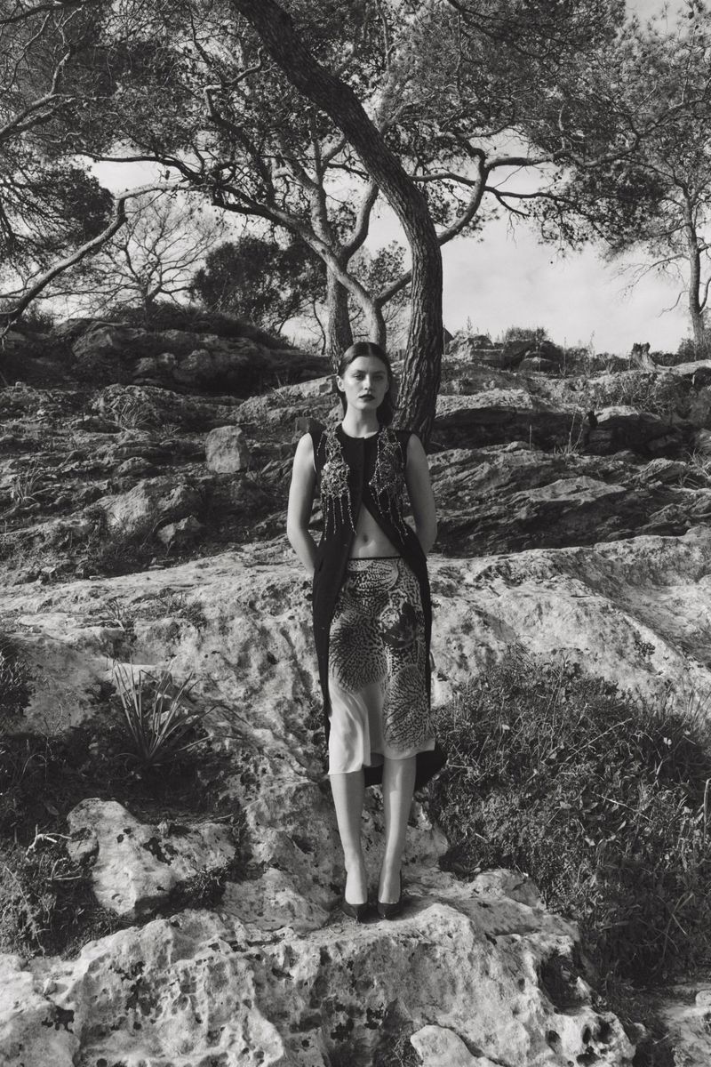 Monochrome fashion portrait of a woman in an embellished vest and patterned skirt, standing on a rocky outcrop with trees in the background.