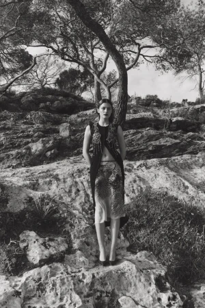 Monochrome fashion portrait of a woman in an embellished vest and patterned skirt, standing on a rocky outcrop with trees in the background.