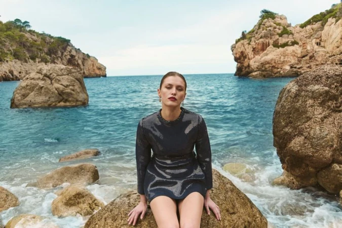 A woman in a dark, shimmering mini-dress sits on a large rock at the ocean's edge, with rocky cliffs and blue water in the background.