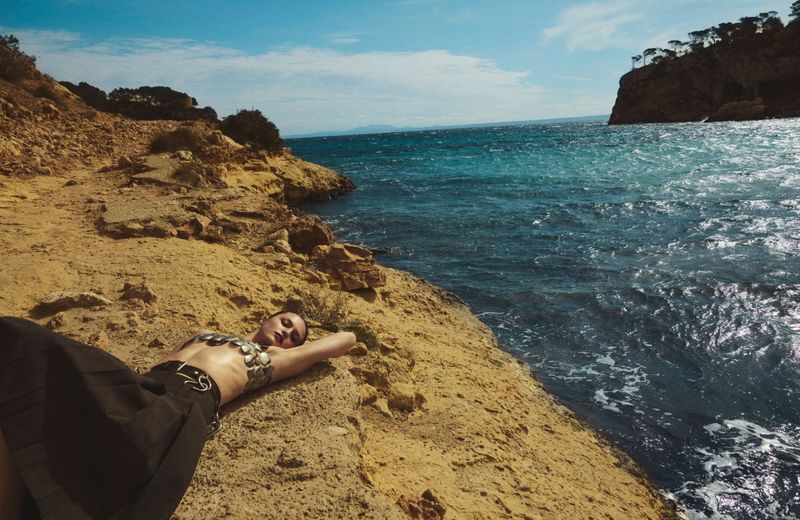 Adult female model with bare midriff lies on a tan rocky coast next to turquoise water on a sunny day, wearing a dark skirt and metal bra.