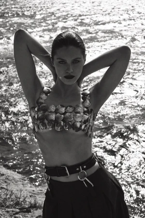 Black and white portrait of a female model wearing a metallic coin-disc top with hands behind her head near water