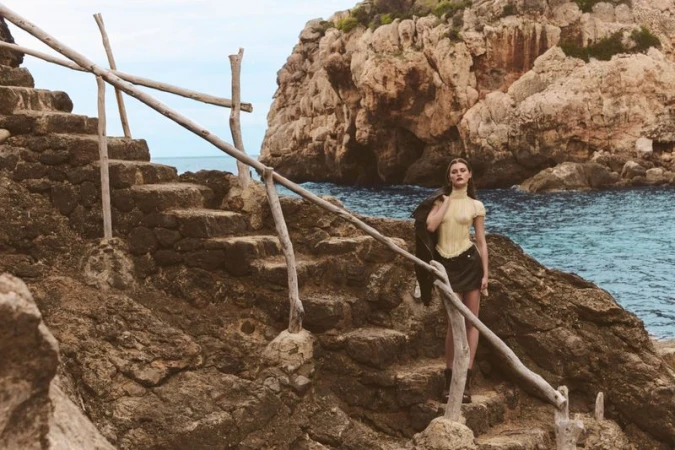 Model standing on stone steps by rocky coast holding a dark jacket for Seduction Magazine Mallorca Wild Trip editorial