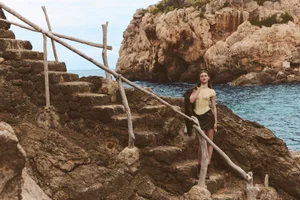Model standing on stone steps by rocky coast holding a dark jacket for Seduction Magazine Mallorca Wild Trip editorial