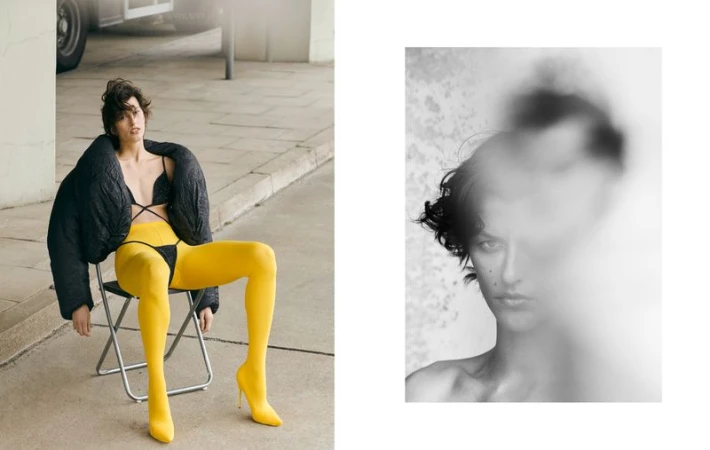 Fashion photo of a woman with short dark hair in a black puffy coat and yellow tights sitting on a chair outside, next to a blurred face.