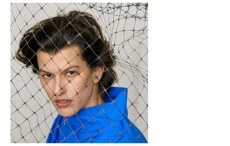 Close-up portrait of a person with dark hair wearing a blue top, partially obscured by a black net in front of a neutral background.
