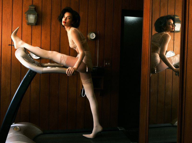 Brunette woman in lingerie strikes a pose on a treadmill inside a room with wood paneling, complemented by a mirrored reflection.