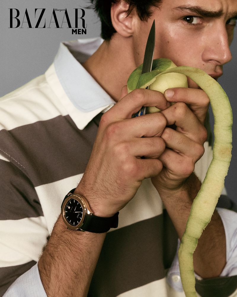 A young adult man peeling a green apple with a knife in a close-up studio shot for a magazine cover, featuring fashion and beauty.