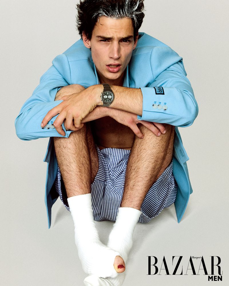 A fair-skinned young adult male fashion model in a blue jacket and striped shorts poses barefoot on a light grey background for a studio shot.