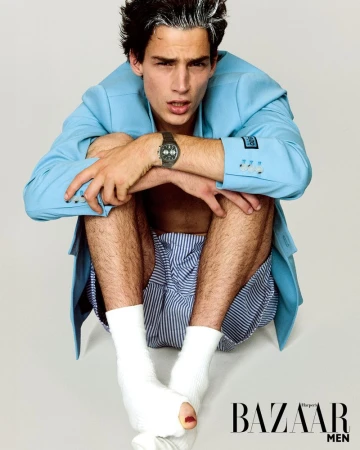 A fair-skinned young adult male fashion model in a blue jacket and striped shorts poses barefoot on a light grey background for a studio shot.