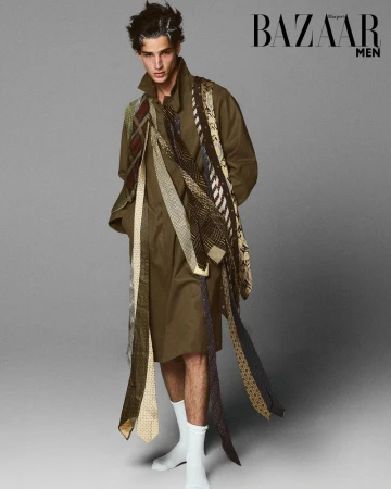 Fashionable young man wearing long olive coat and an array of neckties stands in a photo studio for Bazaar Men editorial shoot.