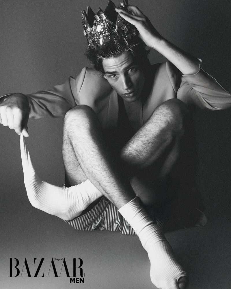 Black and white fashion portrait of a man with a crown wearing white socks and a jacket in a studio environment.