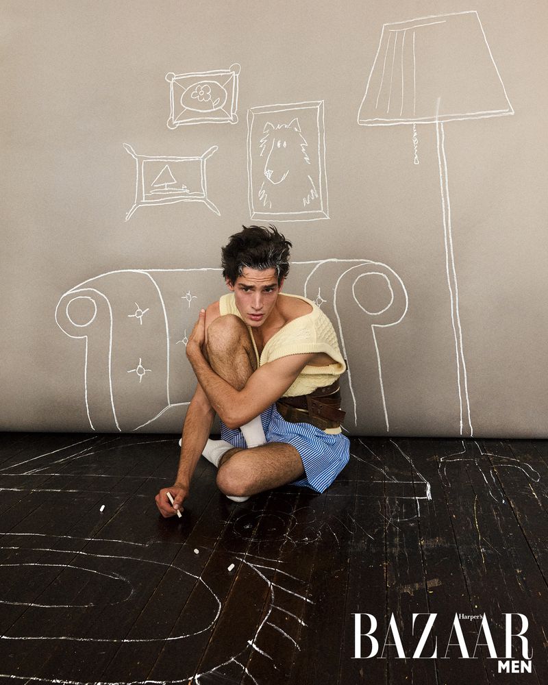 Male model poses on a dark wooden floor in a light-yellow top, blue shorts, and white socks with chalk drawings on the wall and floor.