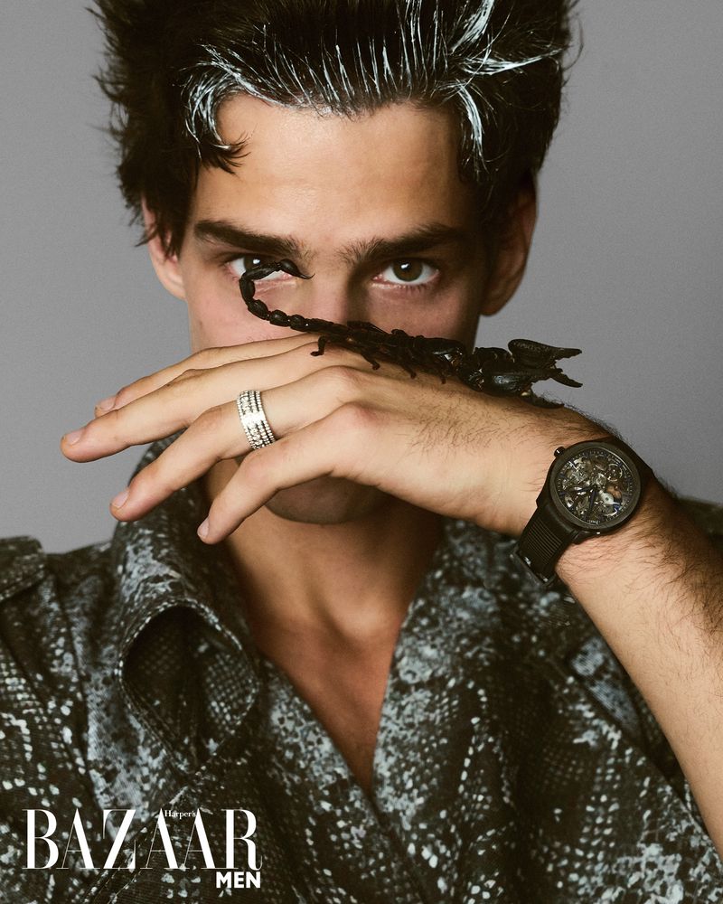 Portrait of a man wearing a printed shirt and ring posing with a black scorpion crawling on his hand in a studio setting.