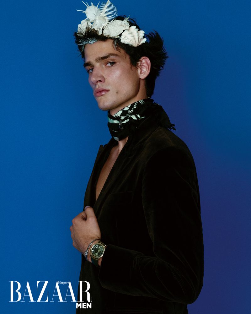 Portrait of a young adult male fashion model wearing a dark jacket, ornate scarf, and feather crown against a solid blue backdrop.