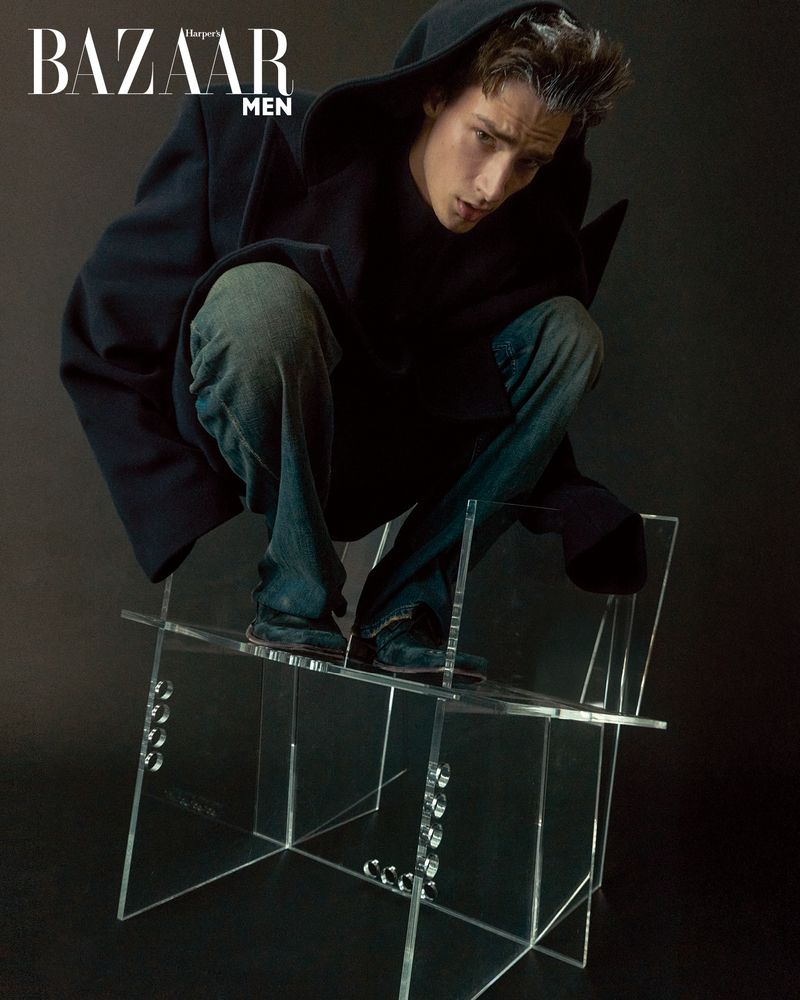 Young man with dark hair sits on clear acrylic chair in dark hooded jacket, blue jeans in studio portrait for Bazaar Men magazine.