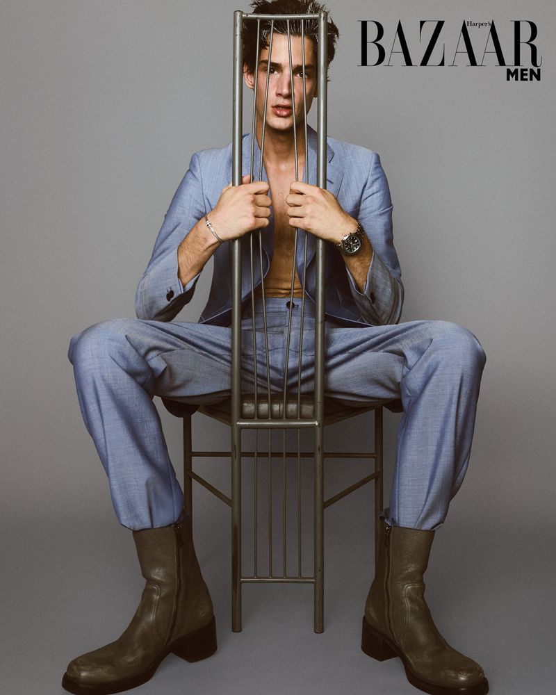 Man in a blue suit poses seated on a chair holding the chair's back, wearing dark boots, with a gray backdrop and the 'Bazaar Men' logo.