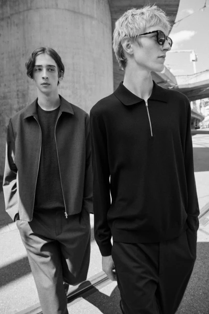 Two adult men pose in modern, dark-colored outerwear and sunglasses on a sunny city street with a concrete overpass. Monochrome photograph.
