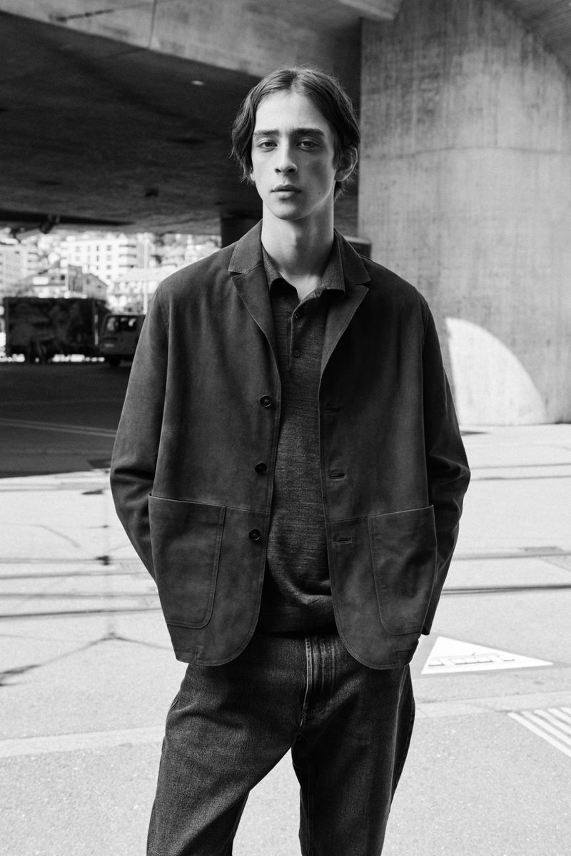 Monochrome portrait of a young adult man in a dark jacket and jeans standing on an urban street under a bridge on a sunny day.