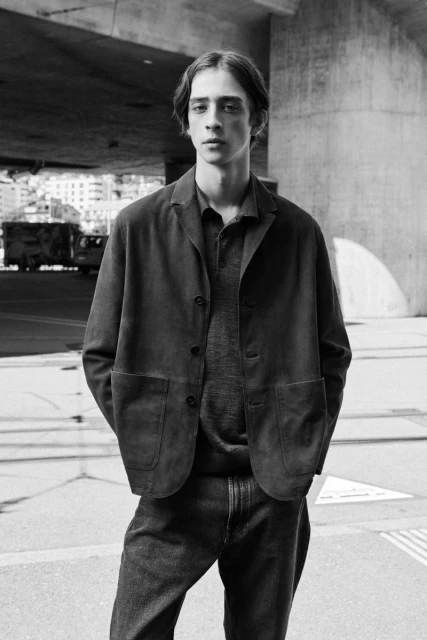Monochrome portrait of a young adult man in a dark jacket and jeans standing on an urban street under a bridge on a sunny day.