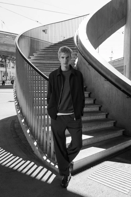 Monochrome portrait of a blond male model in a bomber jacket and trousers standing on a concrete staircase with curved railings.